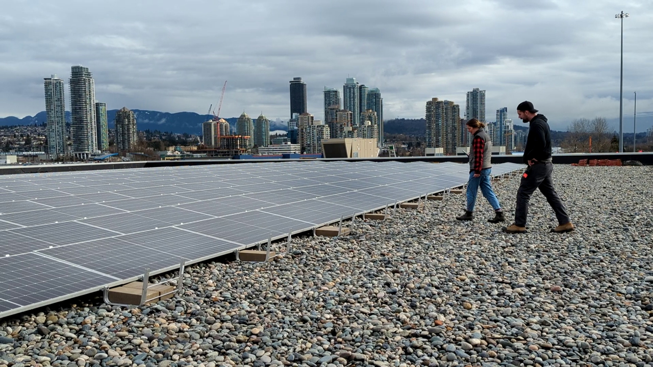 Two people stand in front of a solar array on the roof of a building.