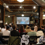 Wide shot from behind of an audience looking at a projector screen.