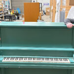 Two people stand either side of a blue coloured standing piano.