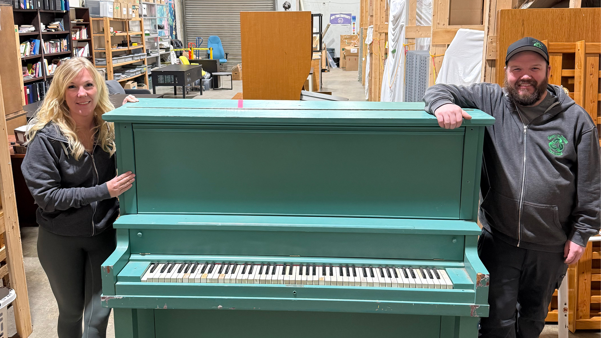 Two people stand either side of a blue coloured standing piano.